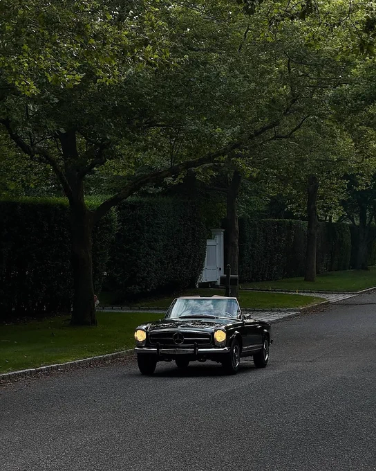 A black vintage Mercedes-Benz 280 SL drives along a tree-lined street with lush green hedges. The car has its headlights on, and the road is quiet with no pedestrians visible.