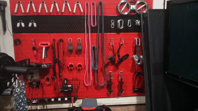 Red Wall Control metal pegboard mounted on a wall, holding neatly organized home gym and workshop equipment including resistance bands, hand grips, carabiners, chains, clamps, cable attachments, and storage baskets, creating a clean, professional, and space-efficient setup.