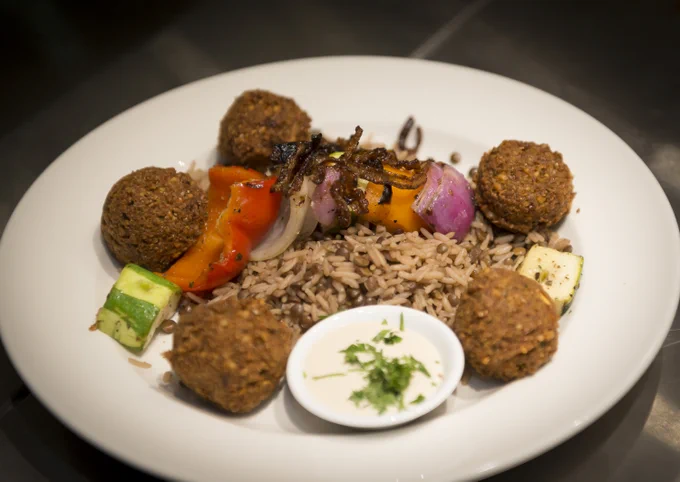 Plate with five falafel balls surrounding a serving of seasoned rice and grilled vegetable skewers, accompanied by a small bowl of creamy herb sauce.