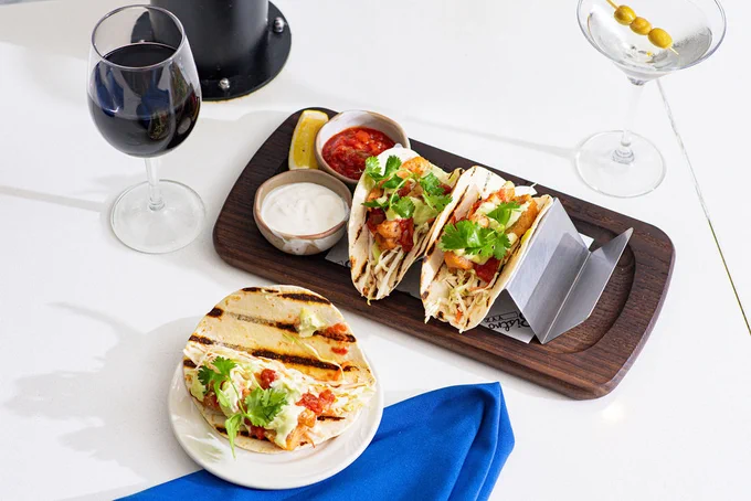 Two chicken tacos with cilantro and sauce on a wooden tray, served with sour cream and salsa, accompanied by a glass of red wine and a martini with olives.