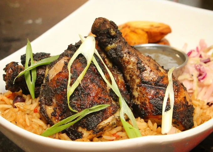 Grilled chicken pieces served over seasoned rice with sliced green onions and a side of sauce and plantains.
