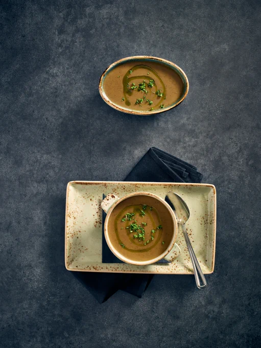 Creamy brown soup garnished with fresh herbs served in a bowl on a rectangular plate with a spoon and dark napkin.