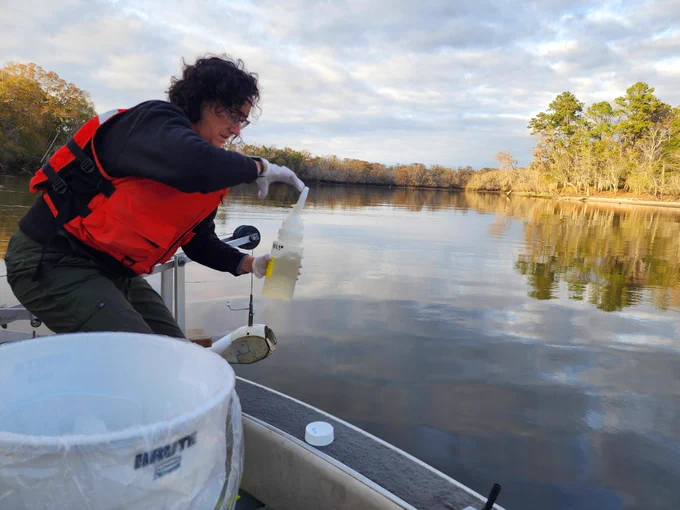 Person in an orange life jacket collecting a water sample from a calm river while on a small boat during daylight.