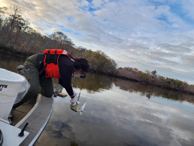 Person in an orange life jacket leaning over a boat to lower a small sampler into calm river water under a cloudy sky.