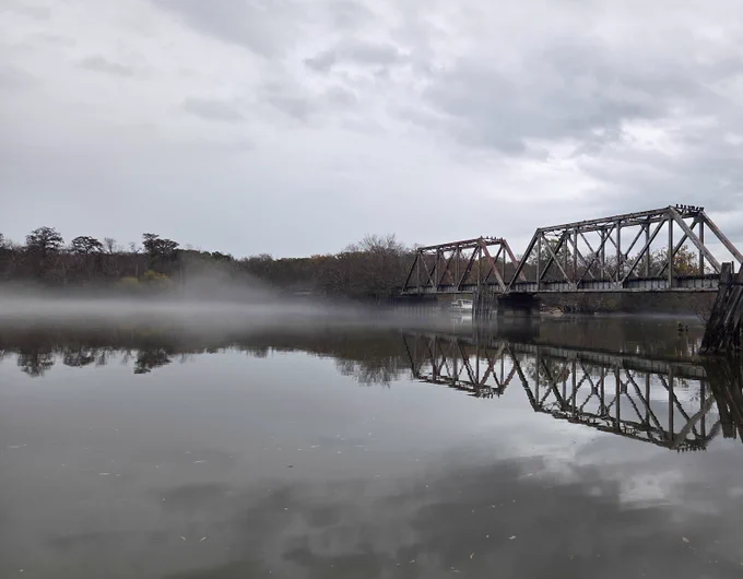 Steel railway bridge spanning a calm river with mist hovering over the water under a cloudy sky.