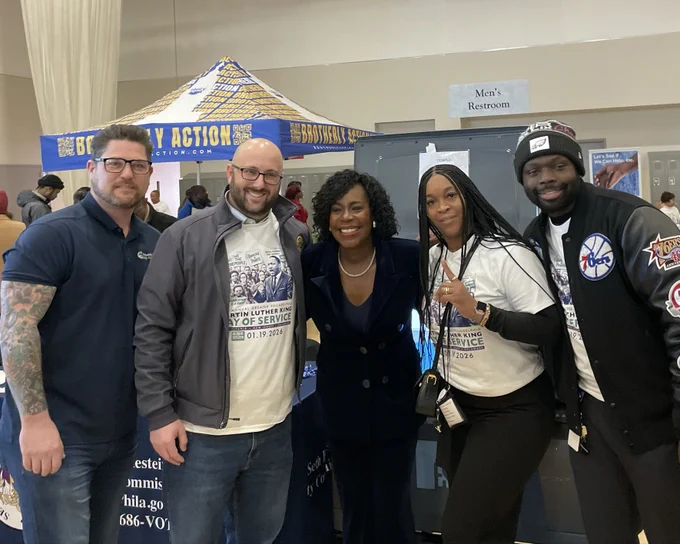 Photo from the Philadelphia City Commissioners showing five people standing together at an indoor community event. Shown are City Commissioner Seth Bluestein, Mayor Cherelle L. Parker, Chairman Sabir’s staff, and additional County Board of Elections staff. Several individuals are wearing white Martin Luther King Jr. Day of Service T-shirts. A blue community outreach tent and informational tables are visible in the background.