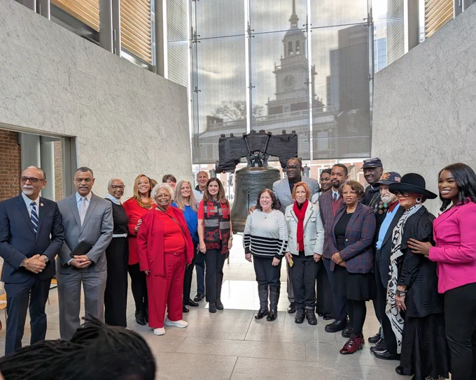 Photo from the Philadelphia City Commissioners showing a large group of elected officials, community leaders, and guests standing together inside the Liberty Bell Center. Vice-Chair Lisa Deeley and City Controller Christy Brady are visible among the group. The Liberty Bell is centered behind them, with large windows revealing Independence Hall in the background.