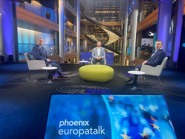 Eine Fernsehstudio-Kulisse mit mehreren Personen, einem gelben ovalen Sitzmöbel und einer großen Bildschirmanzeige mit dem Schriftzug "phoenix europatalks".
