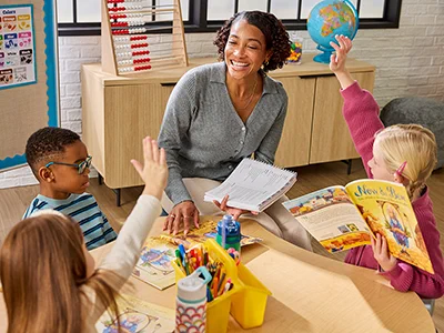 Teacher engaging with young students during a classroom reading activity with colorful books and learning tools.