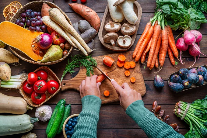 Overhead view of a woman's hands chopping carrots, on a table of fruits and vegetables