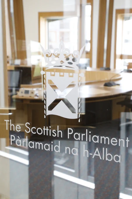 Frosted glass panel with the Scottish Parliament logo and bilingual text reading ‘The Scottish Parliament / Pàrlamaid na h‑Alba’, with a view into a committee room behind it.