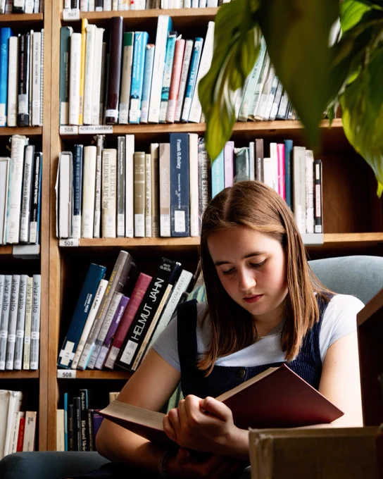 A photograph of a student sitting in a comfortable chair reading a book. They are in a library, and behind them are shelves full of books. 