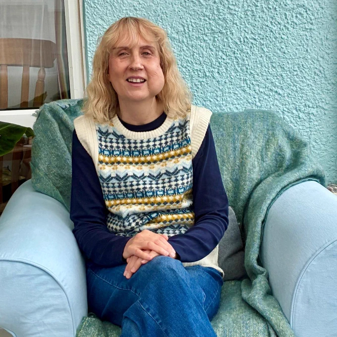 Mary is a white woman in her forties with shoulder-length blonde hair and a fringe. She smiles at the camera, sat on a sofa in a conservatory. She wears a patterned sweater vest, long sleeved top and jeans.