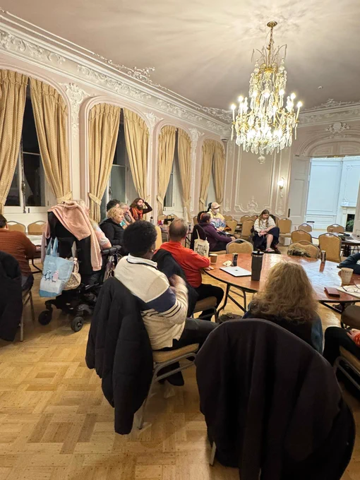 Group of people seated in an ornate room with high ceilings and a large chandelier, engaged in a discussion around tables.