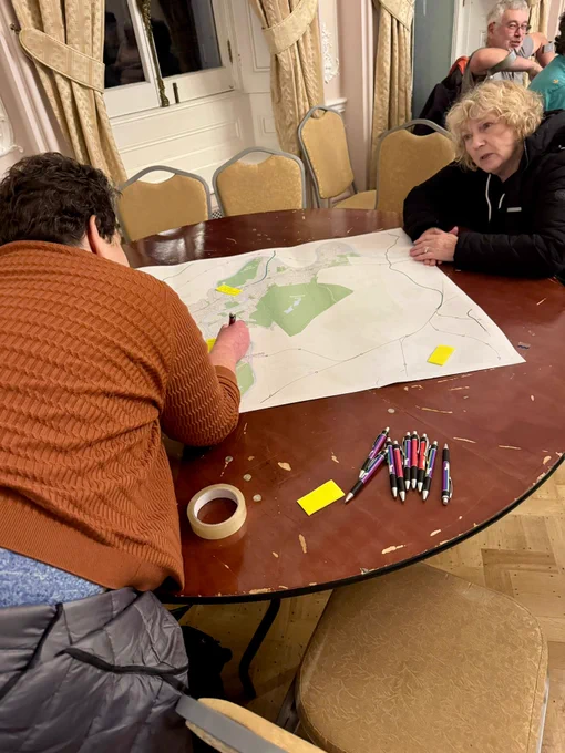 Two people collaborate over a large map spread on a round wooden table in a room with beige curtains and chairs.