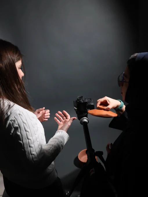 In a dimly lit photography studio, two women are positioned around a camera mounted on a tripod. The background is a solid, dark grey.
On the left, a woman with long brown hair wears a white cable-knit sweater. She is shown in profile, gesturing with her hands as if explaining or directing a shot.
On the right, a second woman is partially visible in silhouette, wearing a dark headscarf and glasses. She holds a small, round terracotta lid or shallow bowl, positioning it carefully in front of the camera lens. A matching terracotta pot sits on the floor below.
The lighting is dramatic, with a strong key light coming from the left that highlights the textures of the white sweater and the women's hands, while leaving the rest of the studio in deep shadow. The composition focuses on the interaction between the two people and the technical process of setting up a shot.