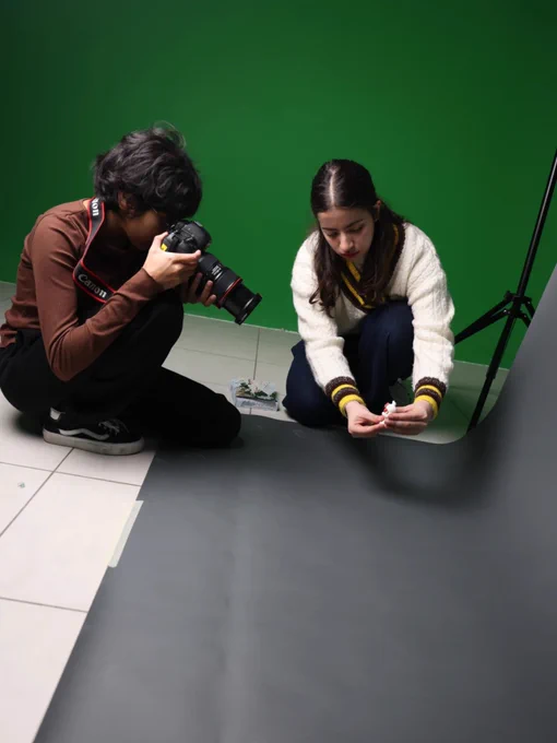 In a professional studio setting with a bright green wall, two young women are collaborating on a product photoshoot.
On the left, a photographer with dark, wavy hair crouches low to the ground. She wears a brown long-sleeved shirt and black pants, holding a black Canon DSLR camera with a large lens close to her face as she focuses on the subject.
To her right, an assistant in a cream-colored cable-knit sweater and dark jeans kneels on the floor. She is carefully adjusting a small white object—which looks like a miniature figure—placed on a seamless grey paper backdrop that curves up from the floor.
The scene captures a moment of quiet concentration. A black tripod stand is visible in the background, and the lighting is bright and even, highlighting the textures of their clothing and the smooth surface of the grey backdrop.