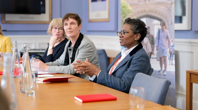 Three women sat at a conference table in a formal meeting room. One person appears to be speaking and is making gestures with her hands.