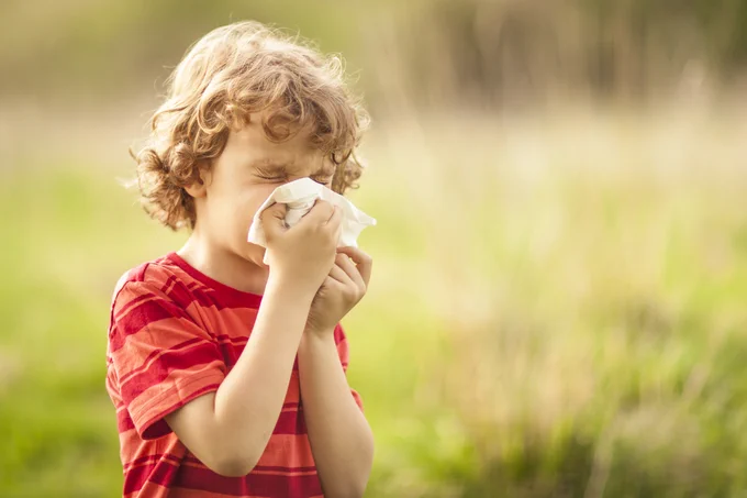 Child in a red shirt blowing nose with a tissue outdoors.
