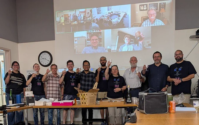Group of people in the room and on video call, rasing a glass to the camera.