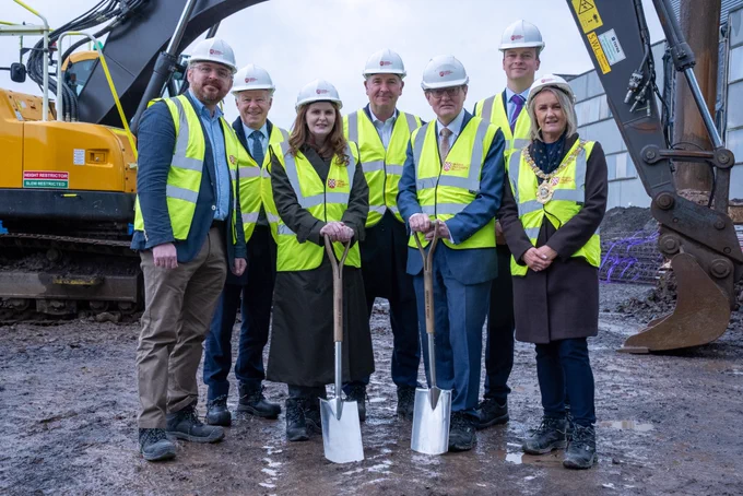 Image shows seven people standing on the building site for the Momentum One Zero expansion, wearing hi-vis jackets and white hard hats. Digging equipment and machinery can be seen in the background behind them.
