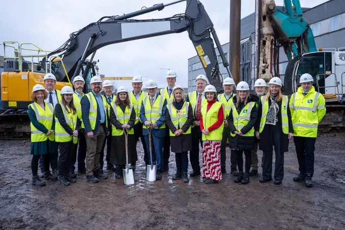 Image shows eighteen people standing on the building site for the Momentum One Zero expansion, wearing hi-vis jackets and white hard hats. Digging equipment and machinery can be seen in the background behind them.
