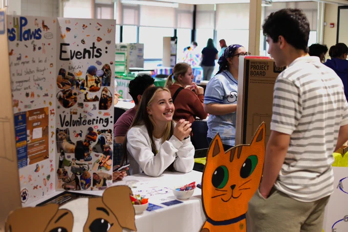 Volunteer at a pet adoption event talks to a visitor beside a display board with photos and info about events and volunteering.
