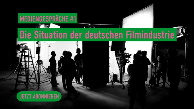 Ein schwarz‑weißes Foto zeigt eine Bühnenszene mit Kameras, Beleuchtungen und mehreren Personen in der Filmproduktion.