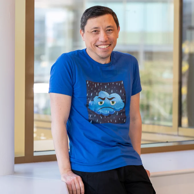 A man in a blue tee shirt with a raincloud graphic sits on a windowsill