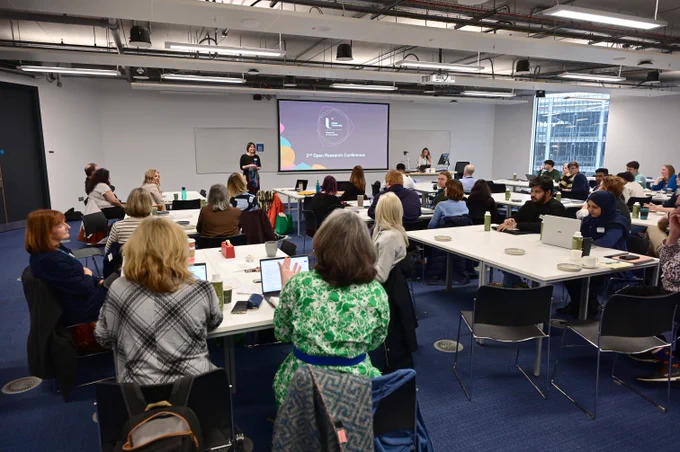 A group attentively listening to a presenter in a modern conference room with a projector screen displaying a presentation.