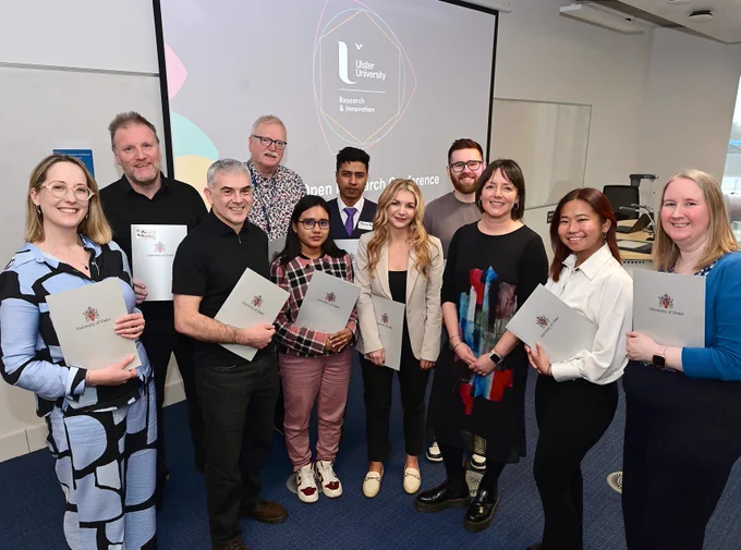 Group holding Ulster University certificates in a classroom setting with a university presentation on screen behind them.