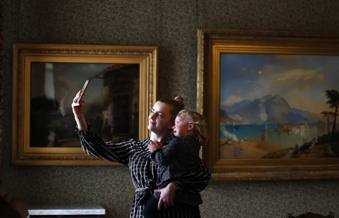 Woman holding a toddler takes a selfie. Two large landscape paintings in gold frames hang in background.