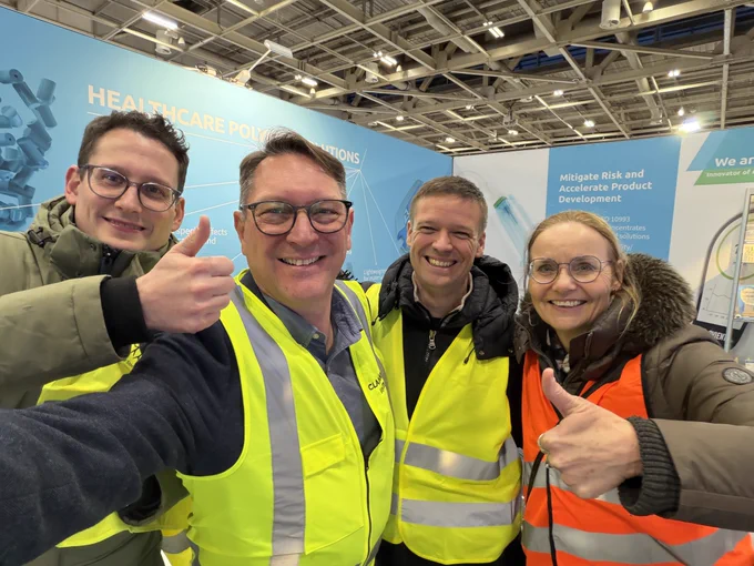 Four people, three men and one woman, smiling in winter coats and high-visibility vests, with two giving thumbs-up, in front of a "Healthcare" banner at a conference.