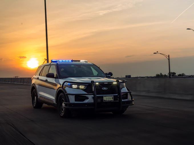 A Ford police SUV with flashing blue lights, driving on a highway at sunset.