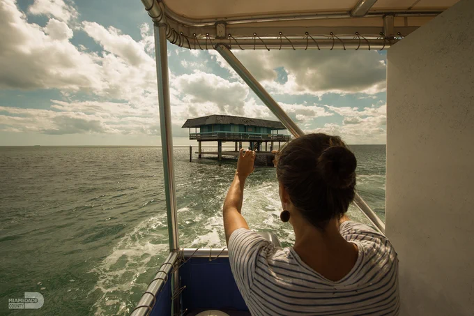 A woman with hair in a bun takes a picture with her phone from a boat. She's facing a structure on stilts in the water, beneath a cloudy sky.