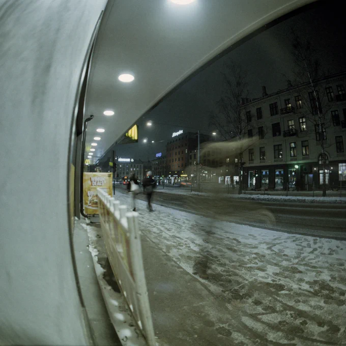 Fisheye photo of people walking on a pavement at night. Light snow on the ground and a street, buildings and lights in the background. The camera was rested against a wall that appears curved due to the fisheye effect.