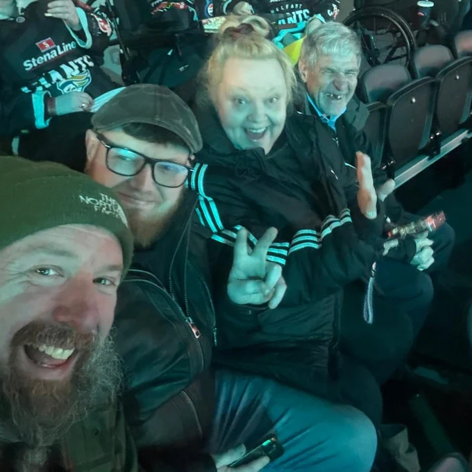 Group of friends sitting in stadium seats, one making a peace sign with fingers, all dressed warmly for an event.