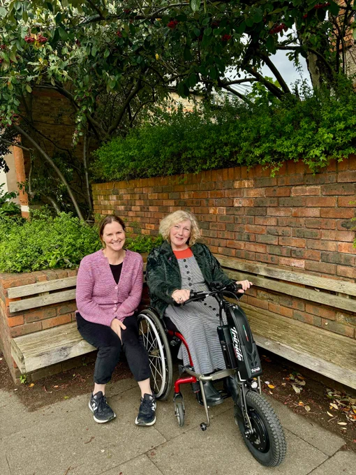 Two women sitting on a wooden bench beside a brick wall, one in a wheelchair with an attached electric scooter, surrounded by greenery.