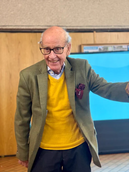 Elderly man wearing a mustard yellow sweater under a green blazer with a patterned pocket square indoors.