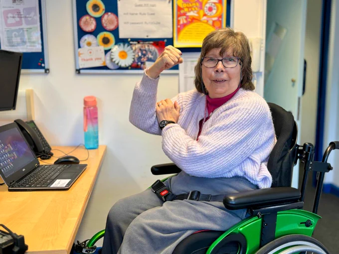 Person in a green wheelchair flexing their arm while sitting at a desk with a laptop and water bottle nearby.