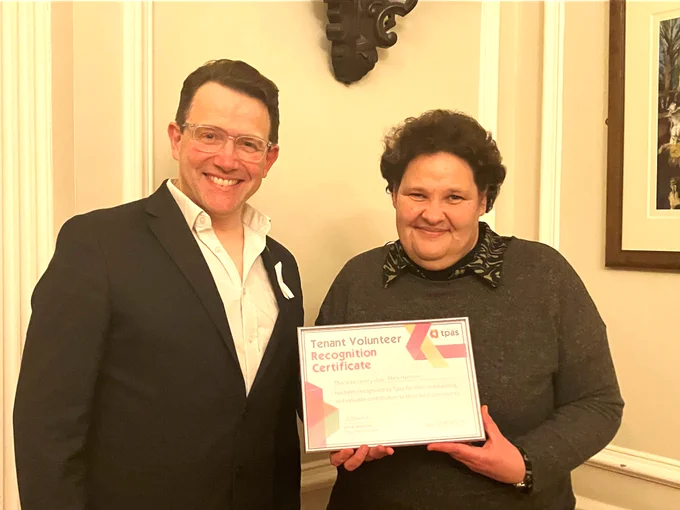 Two people standing indoors, one holding a framed Tenant Volunteer Recognition Certificate from Tpas.