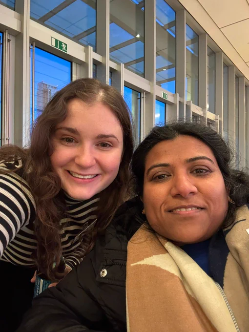Two people taking a selfie inside a modern building with large glass windows and visible exit signs.