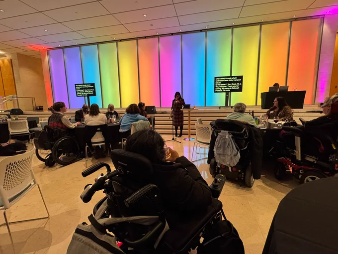 A speaker addresses an audience seated in wheelchairs in a brightly lit room with a colorful gradient wall behind them.