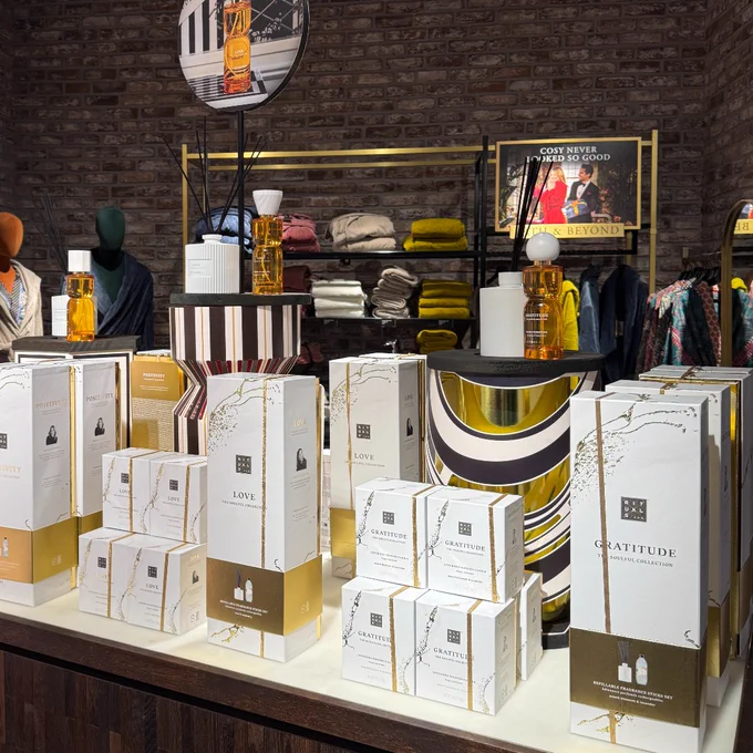 Interior of a modern retail store displaying an assortment of products, including boxed gift items labeled 'Love' and 'Gratitude', arranged on a wooden counter. The background features neatly stacked apparels and accessories under warm lighting.