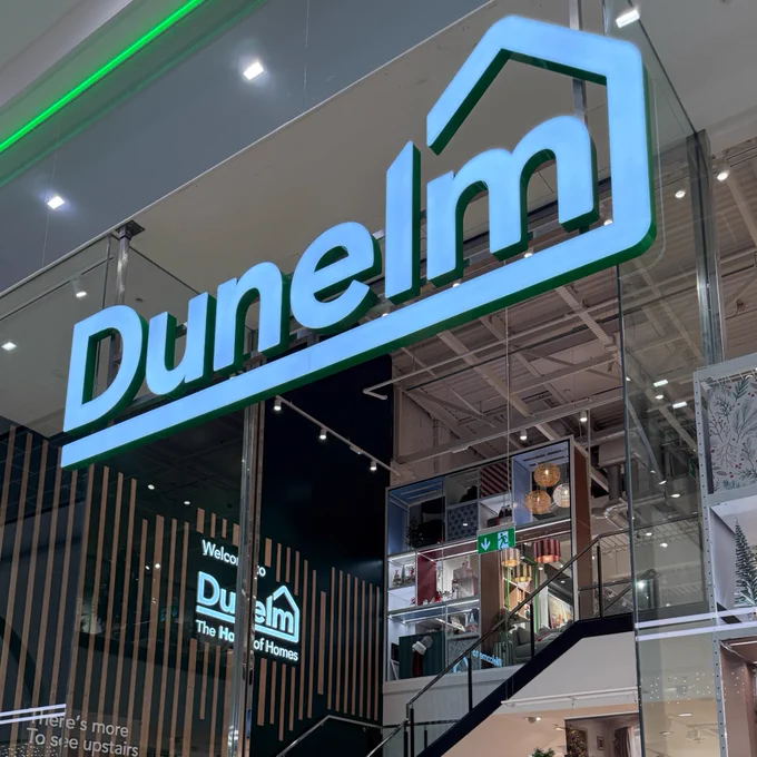Illuminated "Dunelm" store sign above an entrance with visible interior displaying home decor items.