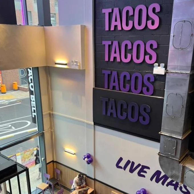 Interior view of a Taco Bell restaurant, showing a sign with "TACOS TACOS TACOS" and "LIVE MÁS" slogans on the wall, viewed from an upper level. One person is seated at a table below.