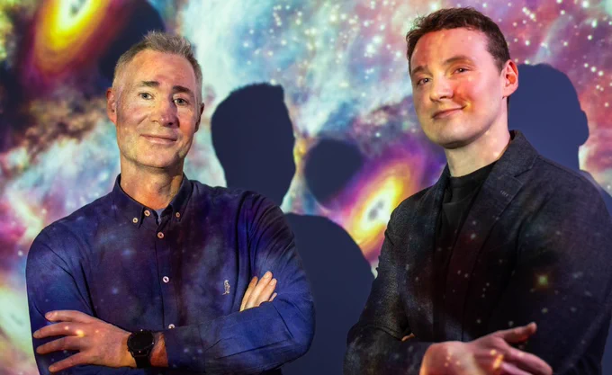 Two men stand confidently with their arms crossed, illuminated by colorful, swirling galaxy projections on their clothing. The vivid cosmic patterns blend with their outfits, creating an ethereal atmosphere against a space-themed backdrop. Both faces are softly shadowed, adding depth to the scene.