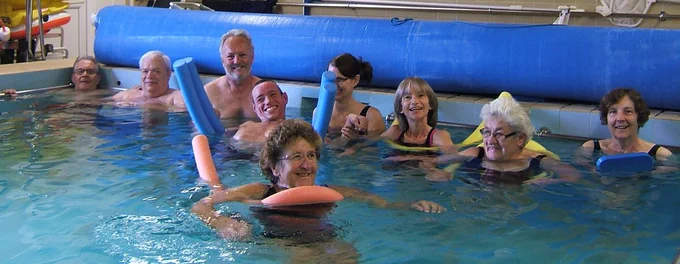 Photo of nine men and woman of all ages in the water of St George's very blue hydrotherapy pool.  They are  laughing and smiling at the camera. 