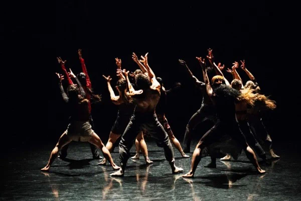 Group of dancers performing a contemporary routine with arms raised against a dark background on stage.
