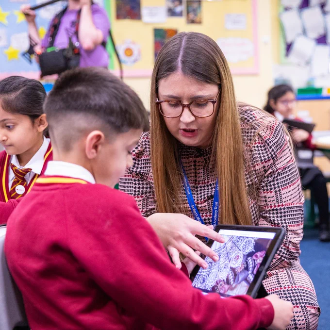 A female teacher kneels down to guide a pupil as they look together at an artwork on an electronic tablet.